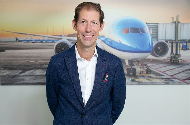 Business-Portrait eines Managers in marineblauem Blazer vor Flugzeug-Kulisse mit Rollfeld und Passagiermaschine im Hintergrund. Das Jackett mit dezentem Einstecktuch unterstreicht die führende Position in der Branche.