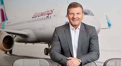 Ein Manager mittleren Alters mit grauem Jackett und weißem Hemd steht über einen Flugsitz gelehnt vor einem unscharfen Hintergrund, der ein Flugzeug der Airline Eurowings zeigt