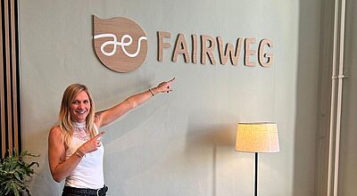 Eine lächelnde Frau mit blondem Haar steht in einem modern eingerichteten Raum und zeigt mit beiden Händen auf ein Wandlogo mit der Aufschrift „FAIRWEG“. Das Logo besteht aus einem stilisierten Blatt mit einem weißen Symbol darin und dem Schriftzug „FAIRWEG“ in Holzoptik. Daneben steht eine Stehlampe, im Vordergrund ist eine grüne Zimmerpflanze sichtbar.