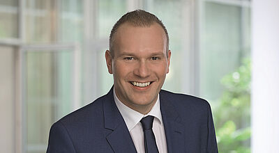 Professionelles Foto eines Managers mit grauem Anzug und weißen Hemd, der in die Kamera lächelt. Die Person ist durch einen unscharfen hellen Hintergrund mit einem Fenster und einem Baum freigestellt.
