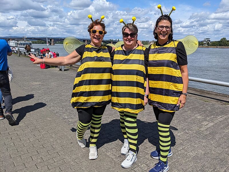 Drei Frauen stehen lachend nebeneinander an einem Uferweg und tragen passende Bienenkostüme mit gelb-schwarzen Streifen, Flügeln und Fühlern mit gelben Pompons. Sie tragen gestreifte Leggings und sportliche Schuhe. Im Hintergrund ist ein Fluss, ein Schiff und eine Brücke zu sehen. Es ist ein sonniger Tag mit teils bewölktem Himmel.