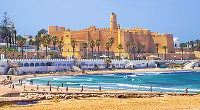 Die historische Festungsanlage Ribat in Monastir mit sandfarbenem Mauerwerk, Palmen, Strand und türkisblauem Meer im Vordergrund.