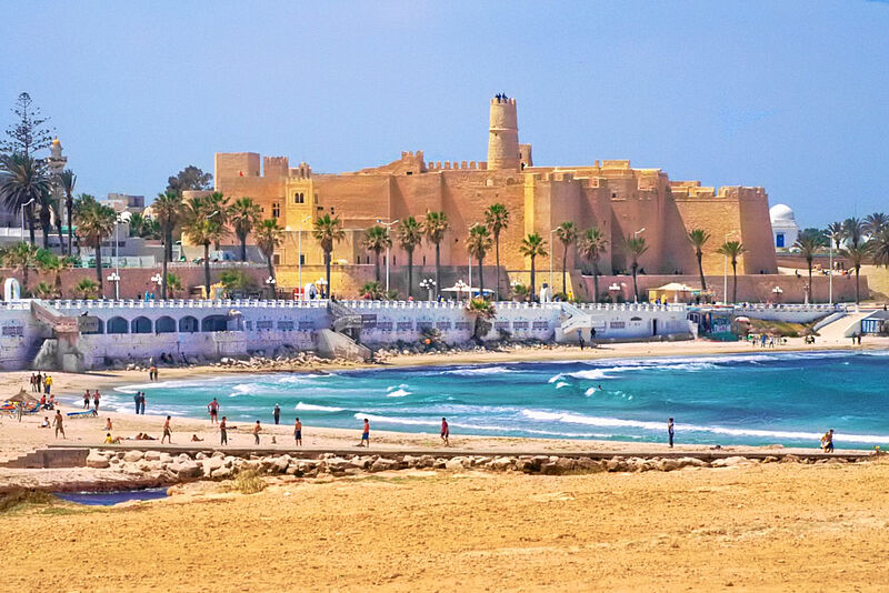 Die historische Festungsanlage Ribat in Monastir mit sandfarbenem Mauerwerk, Palmen, Strand und türkisblauem Meer im Vordergrund.