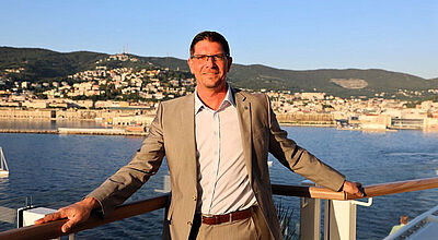 Andreas Nüssel verantwortet künftig die Märkte UK und EMEA An der Reling eines Kreuzfahrtschiffs posiert ein lächelnder Mann mittleren Alters mit brauner Kurzhaar-Frisur und Brille. Er trägt einen hellbraunen Anzug und ein weißes Hemd, im Hintergrund ist eine südeuropäische anmutende Stadt auszumachen.