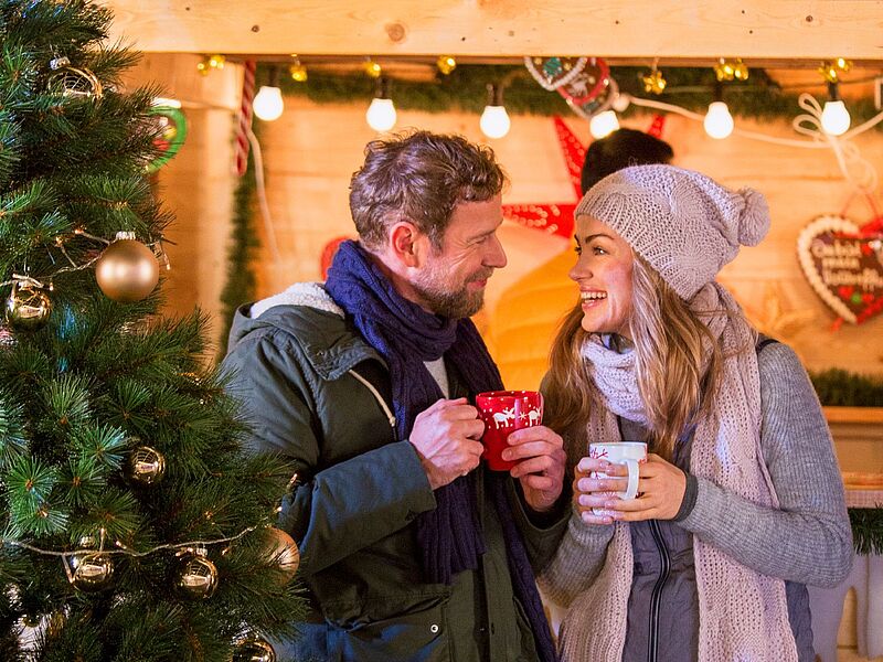 Ein warm angezogenes Paar (Mann und Frau) hält Glühweintassen in der Hand und lächelt sich an. Im Vordergrund ein geschmückter Weihnachtsbaum, im Hintergrund eine Bude aus Holz.