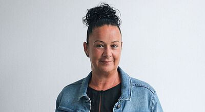 Anke Dreiseidler-Kratz hat beinahe zwei Jahrzehnte Berufserfahrung in der Touristik. Foto: Service-Reisen Frau mit dunklem, hochgestecktem lockigem Haar, trägt eine Jeansjacke über einem schwarzen Oberteil, steht vor einem hellen Hintergrund und lächelt freundlich in die Kamera.