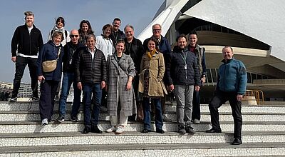 Auf Rundgang in Valencia: der Counterbeirat von Studiosus mit Geschäftsführer Peter-Mario Kubsch (vordere Reihe links) und Vertriebschef Guido Wiegand (vierter von rechts). Foto: Studiosus Eine Gruppe von 15 Personen steht auf einer Treppe vor einem modernen, weißen Gebäude mit geschwungener Architektur. Die Personen tragen Freizeitkleidung, einige mit Jacken und Schals. Sie lächeln in die Kamera, während sie sich für ein Gruppenfoto positionieren. Der Himmel ist blau mit wenigen Wolken, und im Hintergrund sind weitere Gebäude zu sehen.