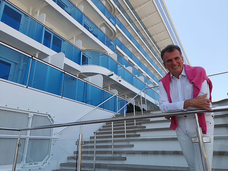 Jörg Rudolph – hier noch zu Costa-Zeiten 2022 an Bord der Toscana – hat Aroya Cruises verlassen. Ein älterer Reederei-Mann mit dunkelbraunem Haar lehnt auf einem Kreuzfahrtschiff an einem Treppengeländer und blickt lächelnd in die Kamera. Er trägt eine weiße Hose und ein weißes Oberhemd und hat einen pinkfarbenen Pullover umgehängt.