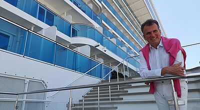 Ein älterer Reederei-Mann mit dunkelbraunem Haar lehnt auf einem Kreuzfahrtschiff an einem Treppengeländer und blickt lächelnd in die Kamera. Er trägt eine weiße Hose und ein weißes Oberhemd und hat einen pinkfarbenen Pullover umgehängt.