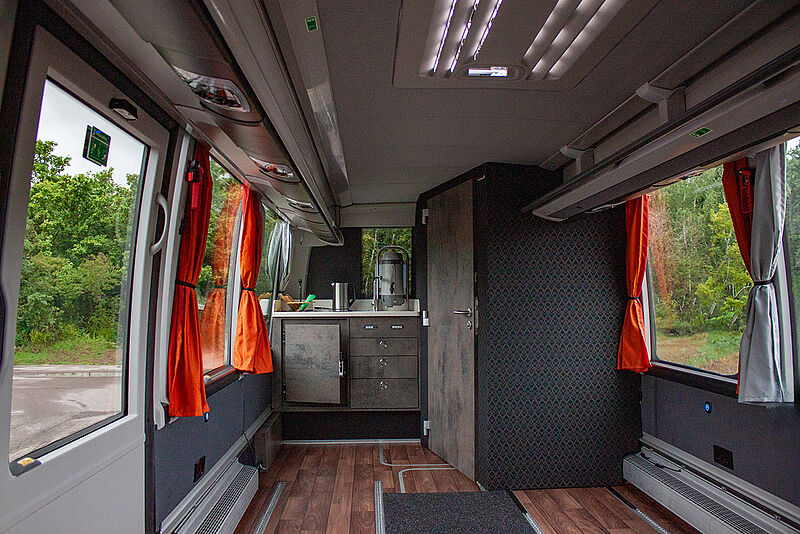 Innenansicht eines modernen, barrierefreien Reisebusses mit wohnlicher Ausstattung. Zu sehen sind eine kleine Küchenzeile mit Spüle, Wasserkocher und Schränken, ein Holzboden, große Fenster mit orange-grauen Vorhängen und eine Tür, die zu einer Toilette führt. Der Bus ist von außen von Grün umgeben.