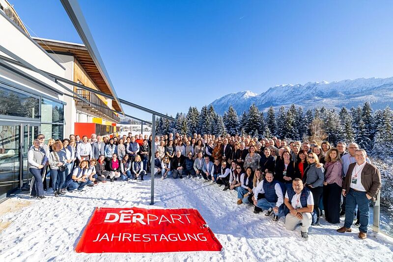 Große Gruppe von Menschen posiert im Schnee vor einem modernen Gebäude mit Blick auf verschneite Tannen und ein Bergpanorama, im Vordergrund liegt ein rotes Banner mit der Aufschrift „DERPART Jahrestagung“.