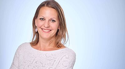 Beate Einhäuser wird zum 1. Juni neue Geschäftsführerin von L’Tur Porträt einer lächelnden Frau mit schulterlangem braunem Haar, die in die Kamera blickt. Sie trägt ein weißes, gemustertes Oberteil und steht vor einem hellblauen Hintergrund.