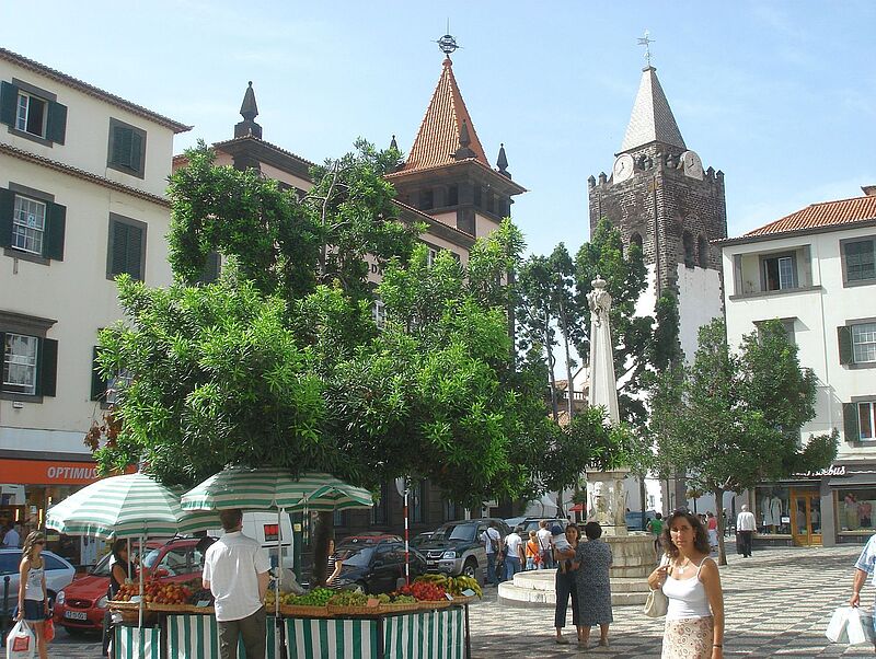 Ein belebter Platz in einer europäischen Stadt mit einer Obst- und Gemüsestandecke im Vordergrund, die von grünen Marktschirmen beschattet wird. Menschen schlendern über den Platz, während im Hintergrund historische Gebäude mit weißen Fassaden, grünen Fensterläden und roten Ziegeldächern zu sehen sind. Ein markanter Kirchturm mit einer Uhr und spitzem Dach ragt in den Himmel, neben ihm steht eine steinerne Statue auf einem Brunnen. Die Atmosphäre wirkt mediterran und einladend.