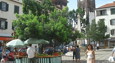 Ein belebter Platz in einer europäischen Stadt mit einer Obst- und Gemüsestandecke im Vordergrund, die von grünen Marktschirmen beschattet wird. Menschen schlendern über den Platz, während im Hintergrund historische Gebäude mit weißen Fassaden, grünen Fensterläden und roten Ziegeldächern zu sehen sind. Ein markanter Kirchturm mit einer Uhr und spitzem Dach ragt in den Himmel, neben ihm steht eine steinerne Statue auf einem Brunnen. Die Atmosphäre wirkt mediterran und einladend.