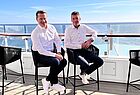 Zwei Männer mit dunklen Hosen und weißem Hemd sitzen auf einem Stuhl und lächeln in die Kamera. Hinter ihnen das blaue Meer. Die Sonne scheint