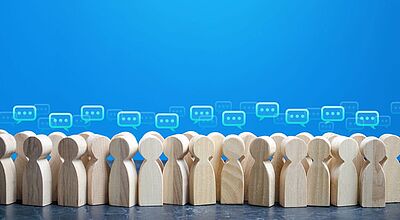 Mehrere Reihen von stilisierten Holzfiguren stehen eng beieinander auf einer dunklen Oberfläche vor einem blauen Hintergrund. Über den Köpfen der Figuren schweben zahlreiche Sprechblasen-Symbole in Grün und Blau, die auf Kommunikation oder Diskussion hinweisen. Das Bild symbolisiert gemeinschaftlichen Austausch, öffentliche Debatte oder kollektive Verhandlungen – möglicherweise im Kontext von Tarifverhandlungen oder Gewerkschaftsaktionen.