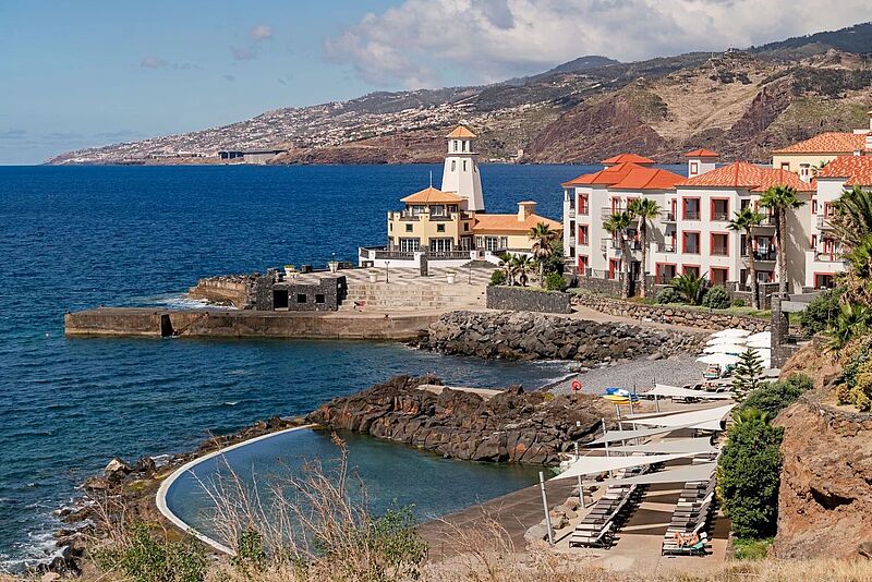 Blick auf ein dreistöckiges Hotelgebäude und einen Leuchtturm. Im Hintergrund Madeira