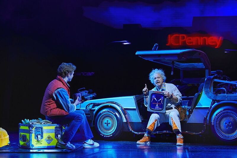 Im März 2026 kommt „Zurück in die Zukunft – Das Musical“ ins Stage Operettenhaus in Hamburg Bühnenszene aus einem Musical: Eine Figur im roten Steppweste sitzt mit einer Videokamera auf einer Kiste, während eine andere mit zerzausten Haaren und weißem Overall vor einem silbernen Auto mit Flügeltüren sitzt und ein leuchtendes technisches Gerät zeigt.
