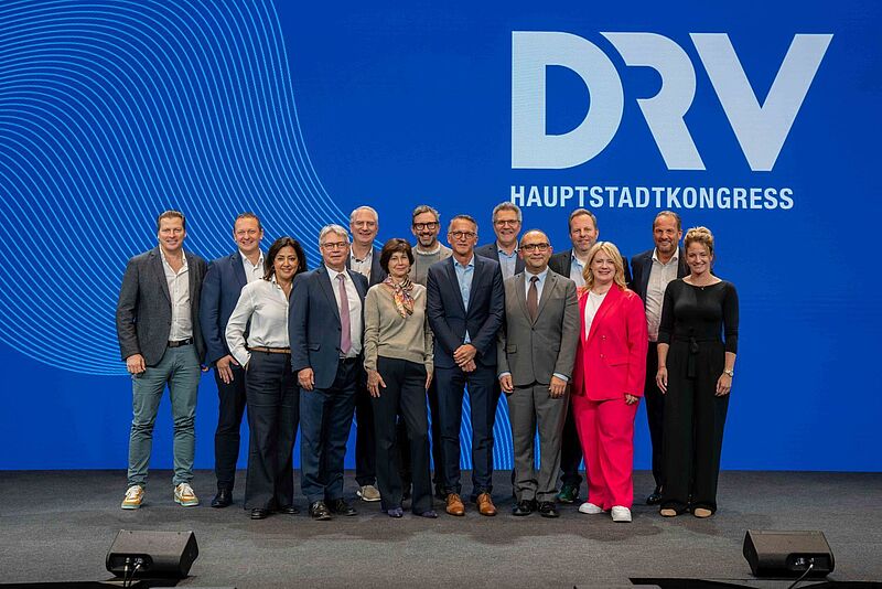 Eine Gruppe von dreizehn Personen steht gemeinsam auf einer Bühne vor einem blauen Hintergrund mit dem Logo „DRV Hauptstadtkongress“. Die Personen sind formell bis halbformell gekleidet und lächeln in die Kamera.