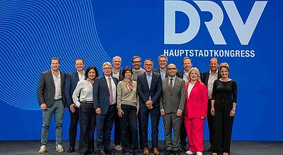 Eine Gruppe von dreizehn Personen steht gemeinsam auf einer Bühne vor einem blauen Hintergrund mit dem Logo „DRV Hauptstadtkongress“. Die Personen sind formell bis halbformell gekleidet und lächeln in die Kamera.
