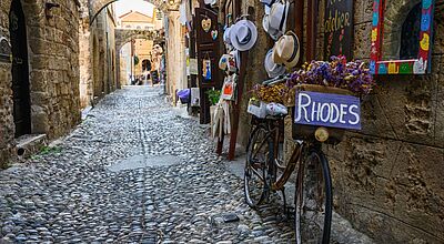 Nach Rhodos führt die Schmetterling-Jahrestagung im kommenden Jahr Enge, historische Kopfsteinpflaster-Gasse in der Altstadt von Rhodos mit alten Steinmauern, kleinen Geschäften und einem dekorierten Fahrrad, dessen Korb mit Blumen gefüllt ist und ein Schild mit der Aufschrift ‚Rhodes‘ trägt.