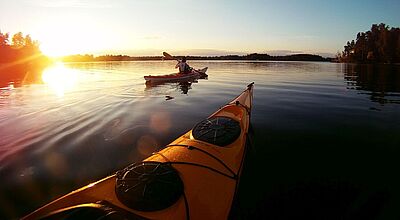Blick über die Spitze eines orangefarbenen Kanus und ruhiges Gewässer auf ein weiteres Kanu, das in den Sonnenuntergang fährt
