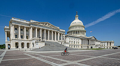 Das Kapitol in der US-Hauptstadt Washington DC vor blauem Himmel an einem sonnigen Sommertag. Der Platz vor dem Gebäude ist leer, lediglich ein Fahrradfahrer fährt durch das Bild.