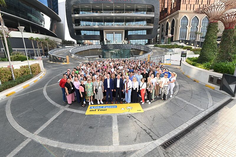 Das Foto zeigt eine Menschenmenge, die sich zur ADAC Jahrestagung vor dem Grand Hyatt Hotel versammelt hat, mit einem gelben ADAC-Reisen-Banner im Vordergrund. Die Teilnehmer stehen auf einer modernen Einfahrt, umgeben von urbanen Bauten, Glasfassaden und exotischen Pflanzen. 
