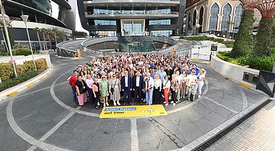 Rund 300 Vertriebsprofis waren zu der Jahrestagung des ADAC Reisevertriebs nach Abu Dhabi gereist. Das Foto zeigt eine Menschenmenge, die sich zur ADAC Jahrestagung vor dem Grand Hyatt Hotel versammelt hat, mit einem gelben ADAC-Reisen-Banner im Vordergrund. Die Teilnehmer stehen auf einer modernen Einfahrt, umgeben von urbanen Bauten, Glasfassaden und exotischen Pflanzen.