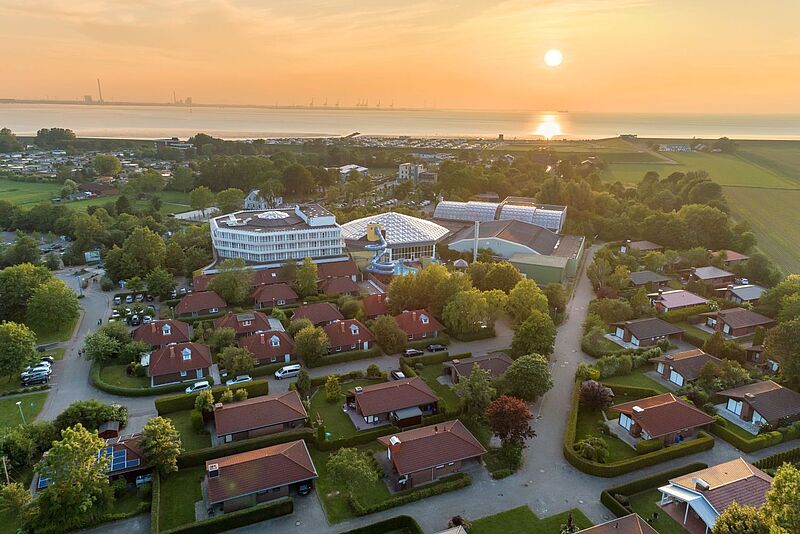 Luftaufnahme eines Ferienparks bei Sonnenuntergang, mit roten Bungalows, einem modernen Hauptgebäude mit runder Architektur und einem Wasserpark mit Rutschen. Im Hintergrund ist die Küste mit einem Deich zu sehen, dahinter das ruhige Meer mit Hafenkränen am Horizont.
