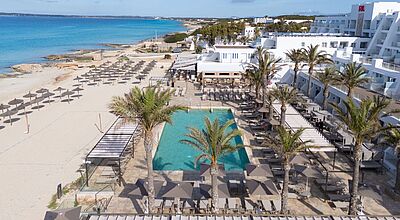 Ein ansprechender weißer Hotelbau an einem hellen feinsandigen Strand mit Liegen, dazwischen ein Pool.