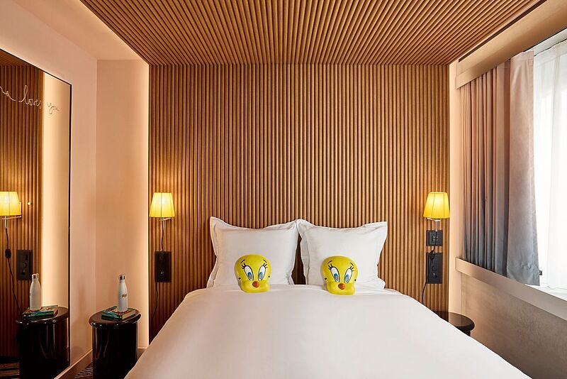 Blick auf ein Hoteldoppelbett mit weißen Laken. Kopfkissen sind im Tweety-Design. Auf dem Nachtisch steht eine Wasserflasse zum Nachfüllen