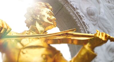 Nahaufnahme einer goldenen Barockstatue eines Geigers mit expressiver Körperhaltung vor hellem Hintergrund mit ornamentalen Verzierungen