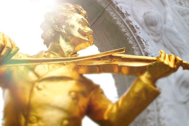 Anlässlich des 200. Geburtstag des Komponisten Johann Strauss hat Mondial das Paket „Meet Johann“ aufgelegt Nahaufnahme einer goldenen Barockstatue eines Geigers mit expressiver Körperhaltung vor hellem Hintergrund mit ornamentalen Verzierungen