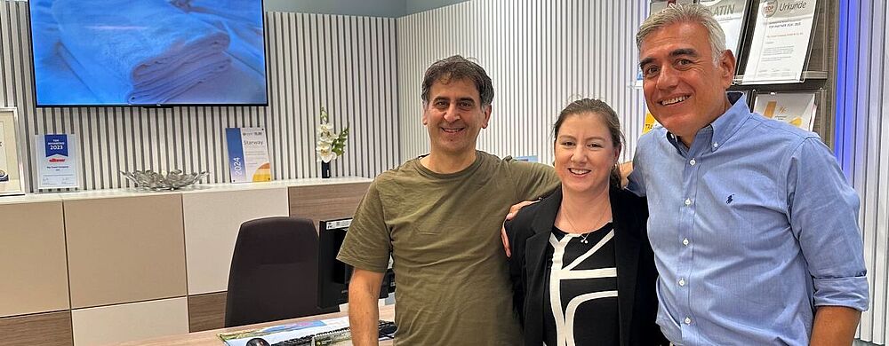 Auf dem Bild stehen drei Personen nebeneinander in einem modernen Büro mit hellen Möbeln und türkisfarbenen Stühlen. Die zwei Männer und eine Frau stehen lächelnd und freundschaftlich Arm in Arm vor einem Schreibtisch. Im Hintergrund sind Auszeichnungen, Urkunden und ein Fernseher mit Handtüchern zu sehen. Zu sehen ist das Reisebüro My Travel Company in der Shopping Mall Skyline Plaza in Frankfurt am Main