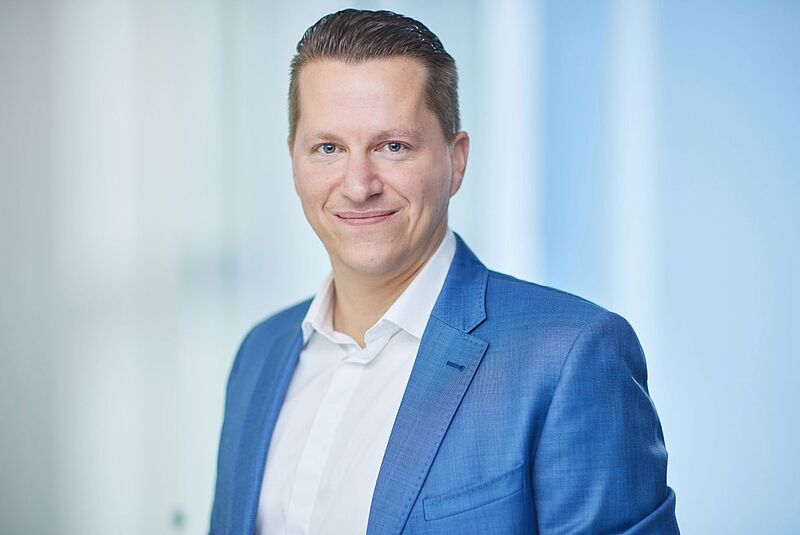 „Ohne Schnickschnack, aber mit Qualität.“ So bezeichnet Benjamin Jacobi, Chef von TUI Deutschland, das neue Konzept von X-TUI Ein lächelnder Mann in einem blauen Anzug mit weißem Hemd steht vor einem hellen, unscharfen Hintergrund. Er hat kurz geschnittene, zurückgekämmte Haare und blickt direkt in die Kamera. Die Atmosphäre wirkt professionell und freundlich.