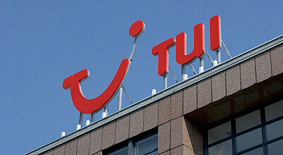TUI prognostiziert weiterhin ein Umsatzwachstum in Höhe von fünf bis zehn Prozent. Foto: TUI Das TUI-Logo in leuchtend roter Schrift auf dem Dach eines Bürogebäudes vor klarem blauen Himmel.