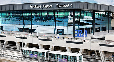 Blick auf das neue Gebäude von Terminal 3 auf dem Frankfurter Flughafen mit großen Glasflächen, im Vordergrund fährt eine Shuttle-Bahn vorbei.