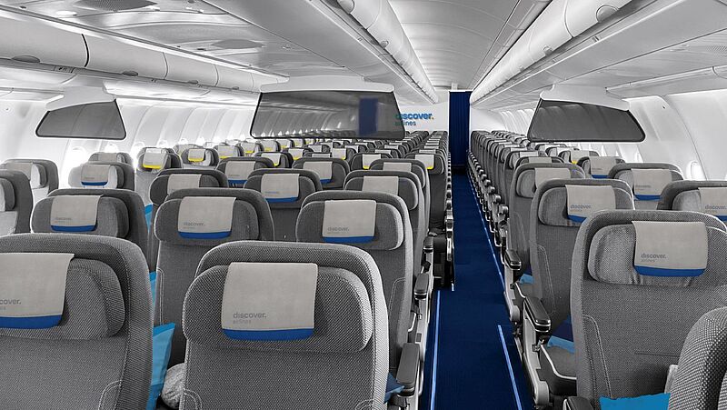 Innenansicht einer Flugzeugkabine von Discover Airlines in der Premium Economy Class mit grauen Sitzen, blauen Akzenten und Kopfstützenbezügen mit dem Airline-Logo, geordnet in mehreren Reihen mit Mittelgang