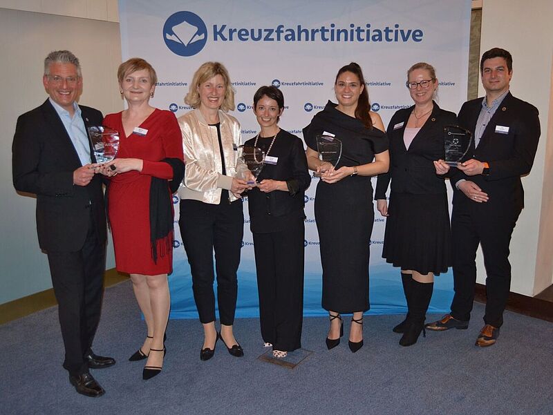 Fünf Frauen und zwei Männer posieren bei einer Preisverleihung mit Awards vor einer Fotowand.