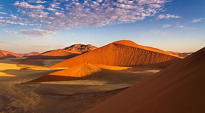 Sonnenbeschienene Sanddünen in warmen Orange- und Rottönen in einer Wüstenlandschaft, mit klarem blauem Himmel und vereinzelten weißen Wolken darüber.
