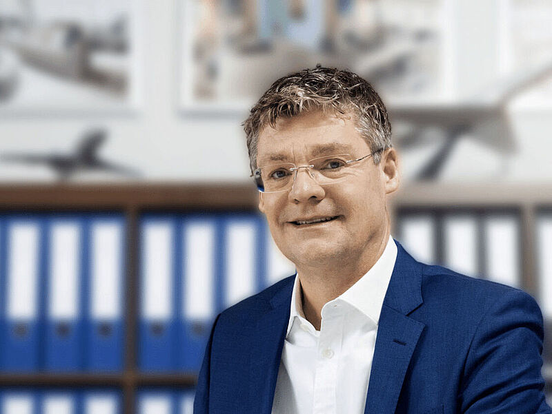 Ein Manager mittleren Alters mit weißem Hemd und blauen Jackett sitzt in einem Büro vor Aktenordnern und Flugzeugmodellen, die im Hintergrund unscharf zu sehen sind