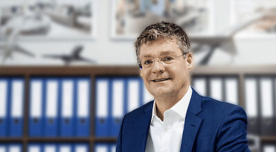 Ein Manager mittleren Alters mit weißem Hemd und blauen Jackett sitzt in einem Büro vor Aktenordnern und Flugzeugmodellen, die im Hintergrund unscharf zu sehen sind