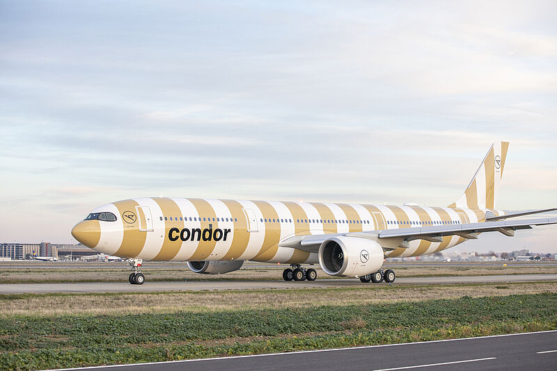 Ein Langstreckenflugzeug der Fluggesellschaft Condor, ein Airbus A330neo, rollt über das Rollfeld. Die Maschine ist in einem auffälligen gold-weißen Streifendesign lackiert. Der Name „condor“ ist in schwarzer Schrift auf dem Rumpf zu sehen, das Unternehmenslogo befindet sich an Triebwerken und Seitenleitwerk. Der Himmel ist leicht bewölkt, im Hintergrund sind Gebäude und weitere Flugzeuge zu erkennen.