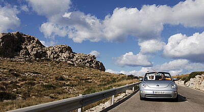 Silbernes Cabrio auf Bergstraße, felsige Landschaft und blauer Himmel mit weißen Wolken im Hintergrund.