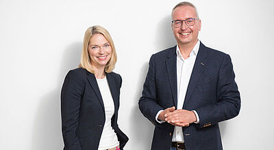 Eine Managerin und ein Manager in legerer Business-Kleidung stehen lächelnd vor einer weißen Wand.