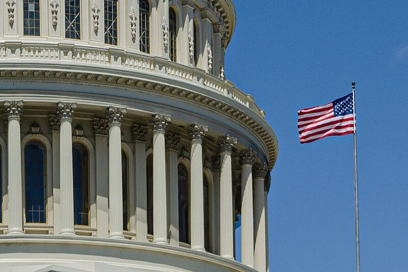 Brand USA wirbt weltweit für Reisen in die Vereinigten Staaten – künftig vielleicht wieder mit mehr Budget Zu sehen ist auf der linken Seite ein Teil-Ausschnitt des Kapitols, eine mit Säulen geschmückte Kuppel, auf der rechten Seite weht die US-Flagge vor blauem Himmel.