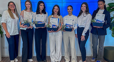 Eine Gruppe von sieben jungen Frauen steht lächelnd nebeneinander in einem hellen Raum vor einem großen Bildschirm mit der Aufschrift „Stiefvater – Herzlich Willkommen zum Ausbildungsstart 2025“. Fünf der Frauen halten Geschenkkörbe mit Firmenunterlagen, Snacks und kleinen Pflanzen in den Händen. Alle sind gepflegt gekleidet, teils in Business- oder Business-Casual-Outfits.