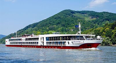 Die Bellejour ist ab 2026 für Nicko Cruises auf der Donau unterwegs Ein zweistöckiges Flusskreuzfahrtschiff in der Lackierung weiß und rot fährt auf einem Fluss. Im Hintergrund ein grüner Berghang.