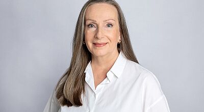 Porträtfoto einer älteren Dame mit langen, grau-braunen Haaren, die lächelnd in die Kamera schaut. Der Studiohintergrund ist grau.